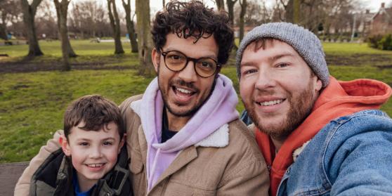Two adults and a young child are taking a selfie in a park. All are smiling happily.