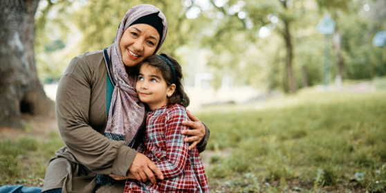 A smiling adult wearing a headscarf sits in parkland. In their arms they are cuddling a child who is looking upwards with a slight smile.