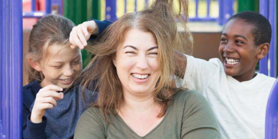 An adult sits on a climbing frame in a playground between two children, who are playing with the adult’s hair. They are all laughing.